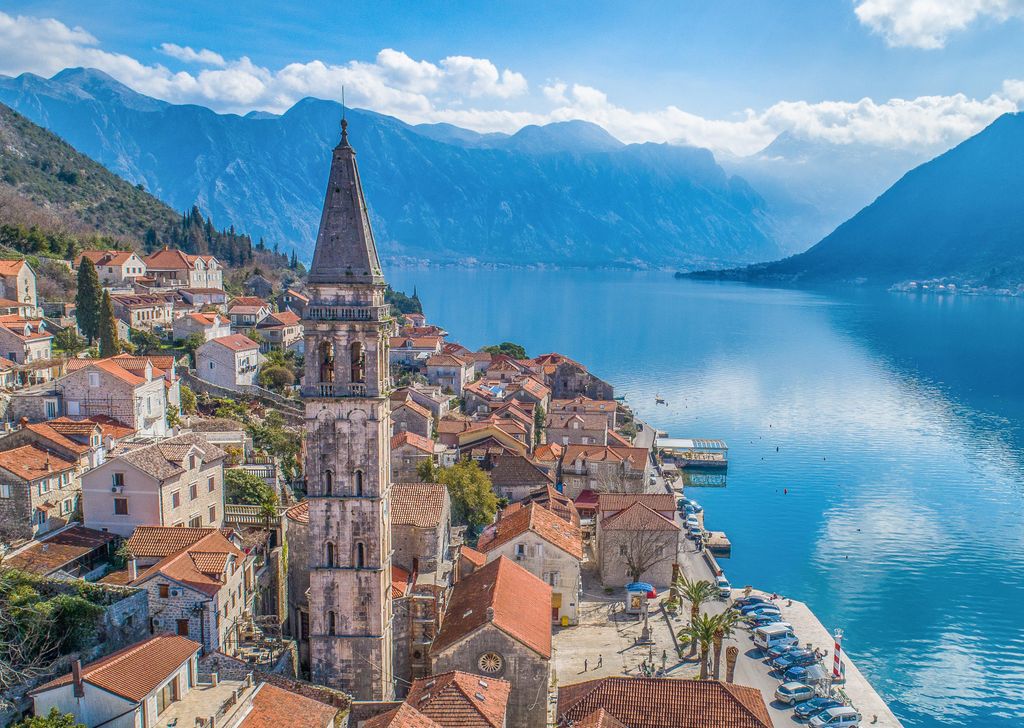 Une vue aérienne de Perast, le Monténégro, montre un clocher en pierre historique s'élevant au-dessus des toits de bâtiments carrelés rouges regroupés le long de la baie bleu vif de Kotor. Les montagnes majestueuses encadrent l'eau en arrière-plan.