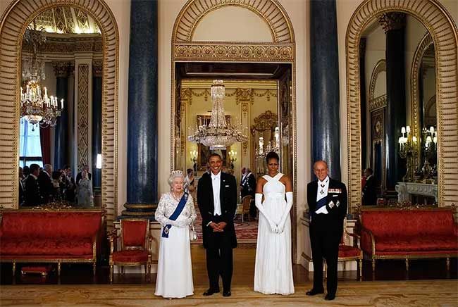 the late queen elizabeth and prince philip stand alongside president obama and his wife michelle in front of a high arch between red velvet covered seats