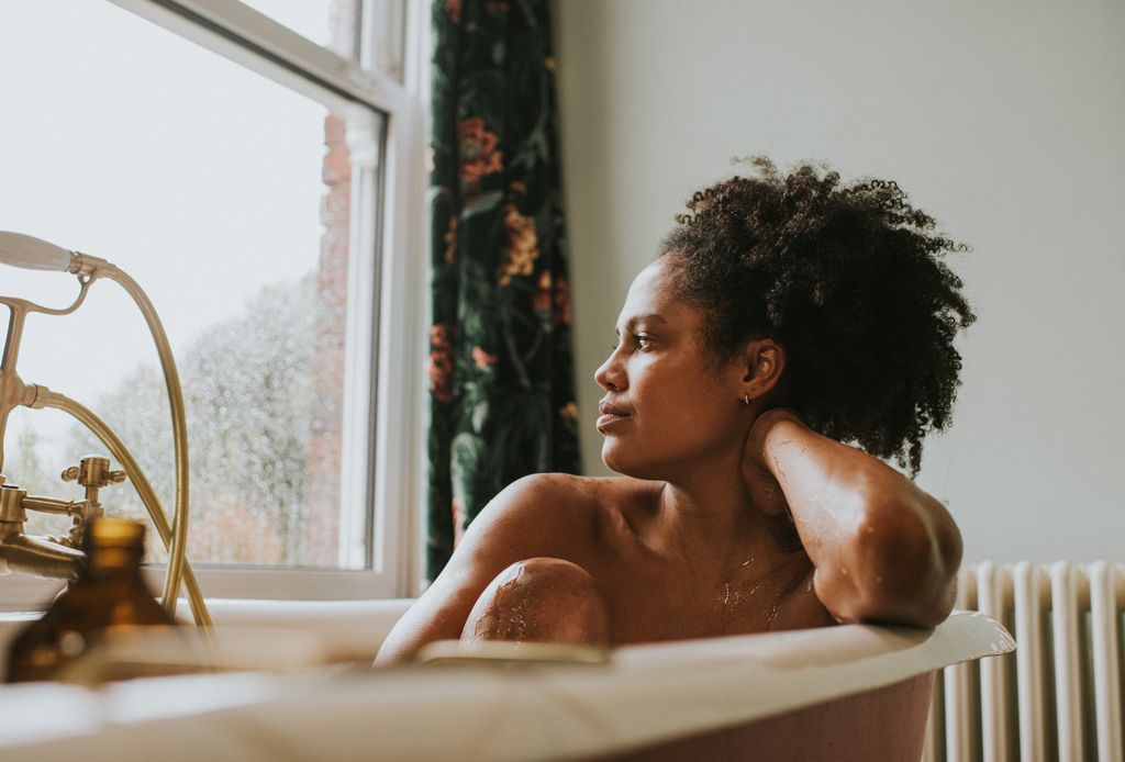 A beautiful woman bathes in a roll top bath. She seems distracted as she rests her head on her arm and gazes out the window. Space for copy.