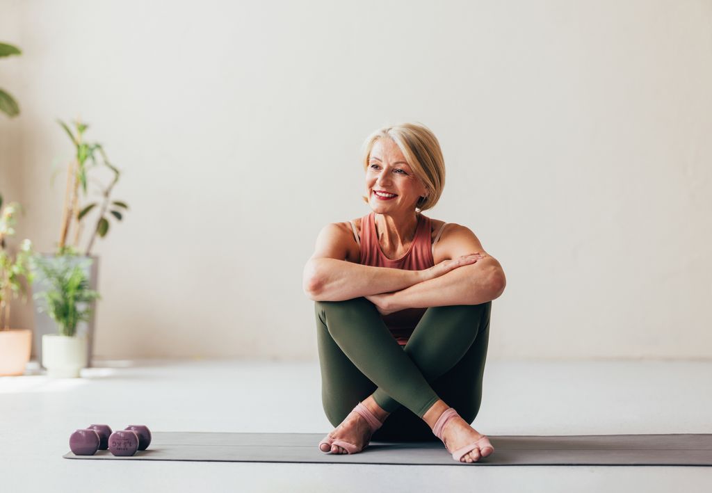 A cheerful senior woman seated on a yoga mat at home, enjoying a relaxing moment after exercising with weights.