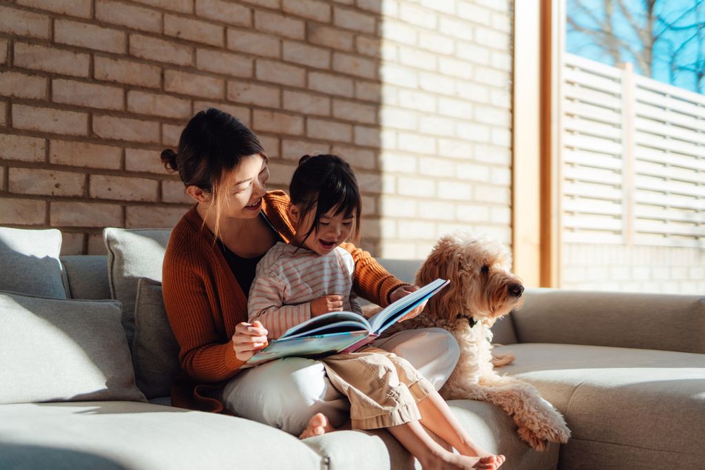 Young Asian mother reading picture book for cute daughter while sitting with their family dog on sofa at home