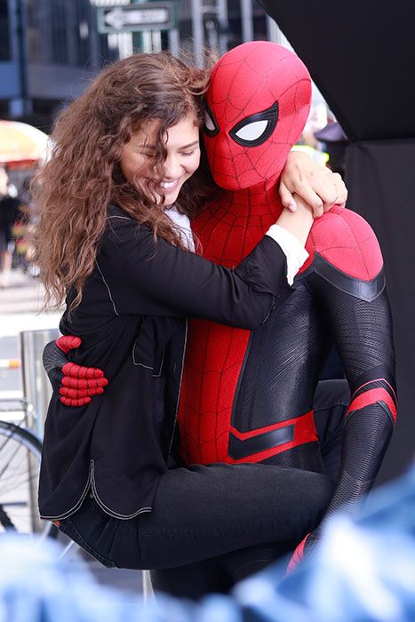 Zendaya holding on to a Spider-Man suited Tom Holland, smiling on set
