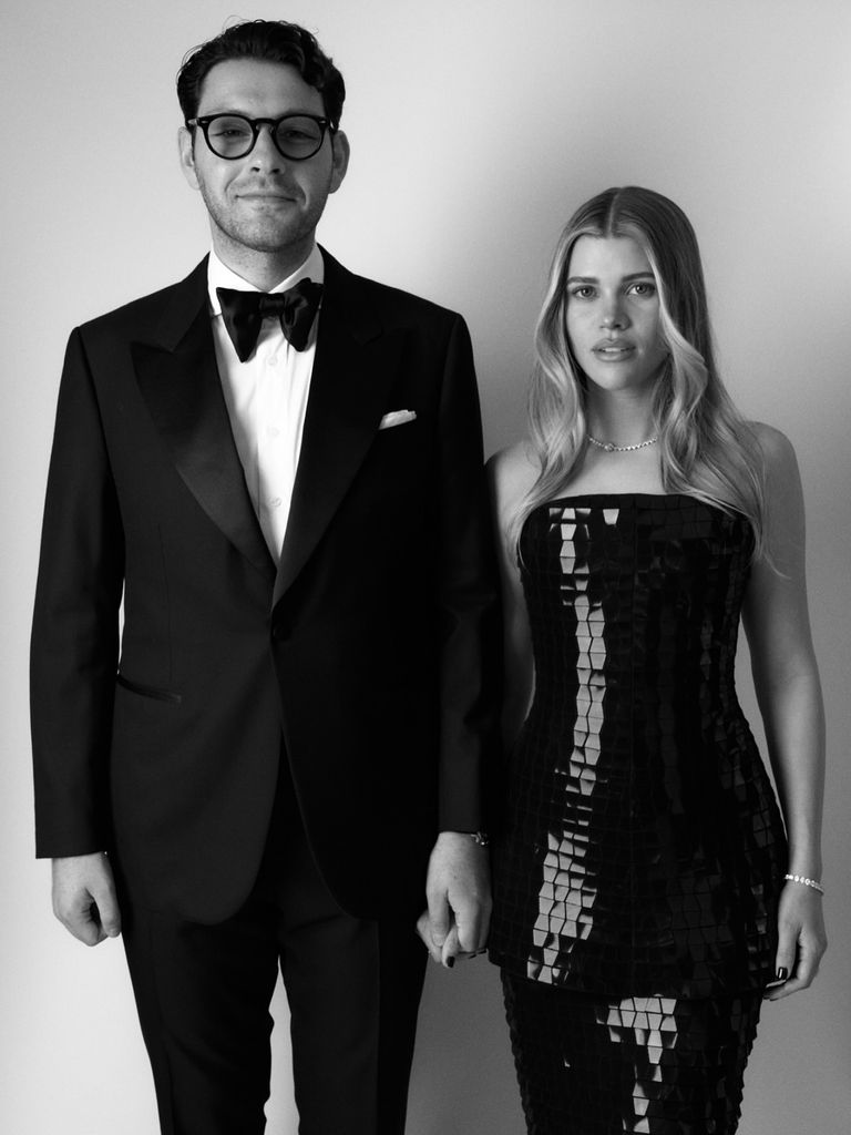 Sofia Richie and her husband Elliot Grainge stand side by side in a black and white picture. He is wearing a black suit and bow tie and she is wearing a strapless sequin black dress