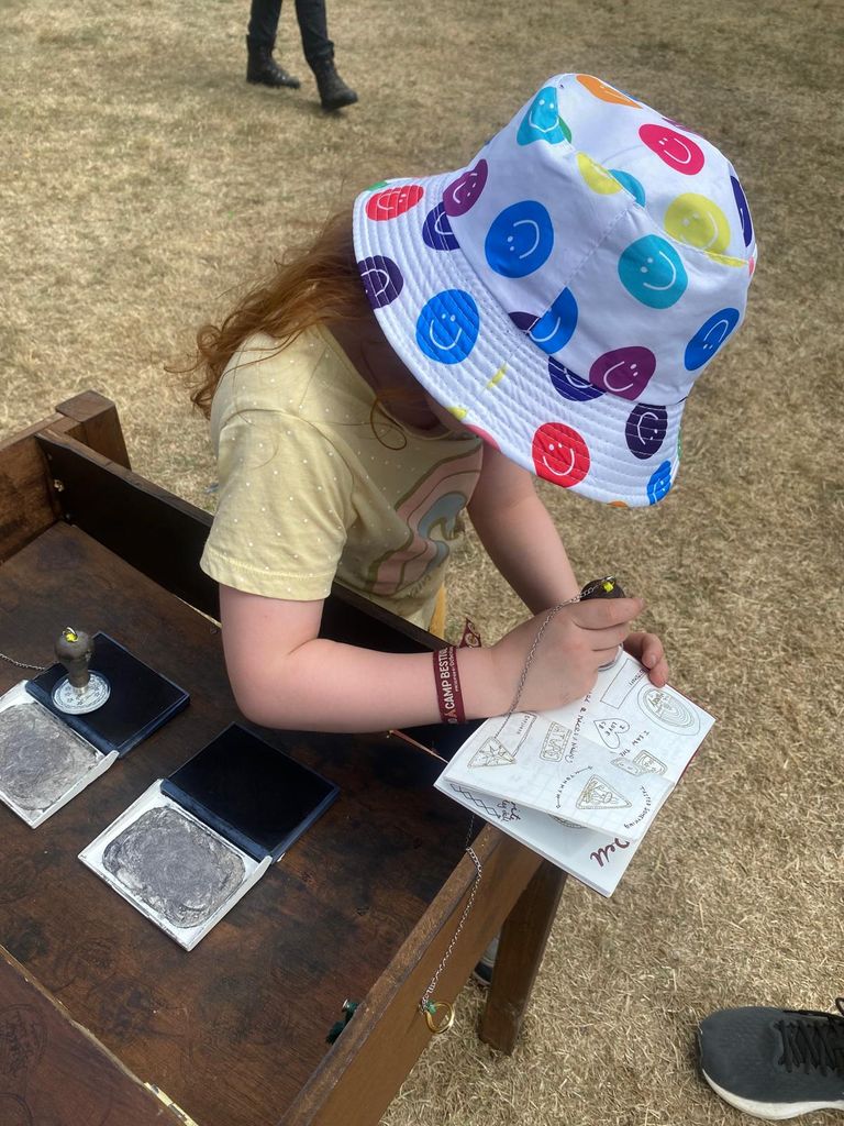 Little girl outside wearing a colourful bucket hat and yellow t-shirt leaning on a desk stamping a book 