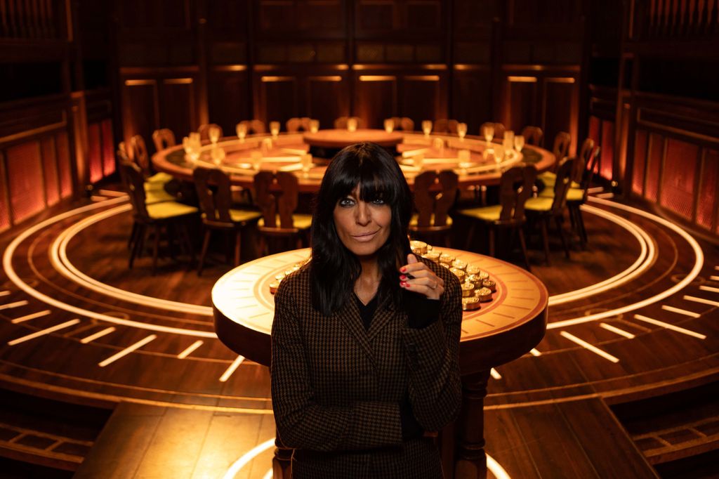 Claudia Winkleman in tweed jakcet stood in front of round wooden table