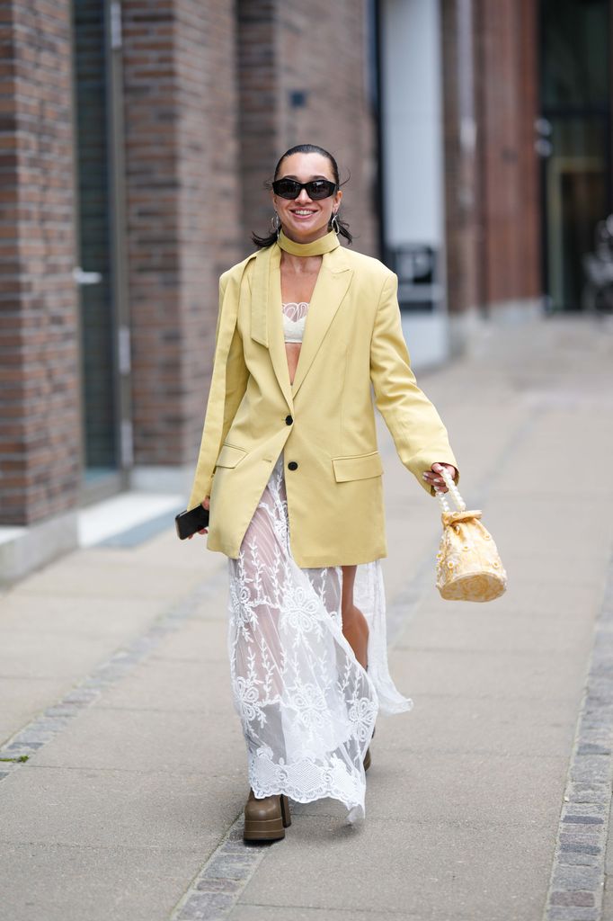  guest wears black sunglasses, a pale yellow long blazer jacket, silver earrings, a white latte tulle lace pattern shoulder-off / cropped top, a matching white lace print pattern tulle slit / split long skirt, dark brown shiny leather platform soles block heels ankle boots , outside Lovechild 1979