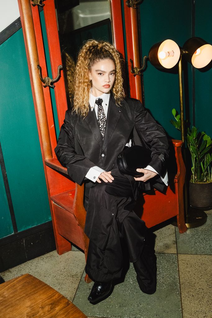 Kiana Alexis in oversized black suit with white shirt and patterned tie leans against red mirror at Met Gala after-party.