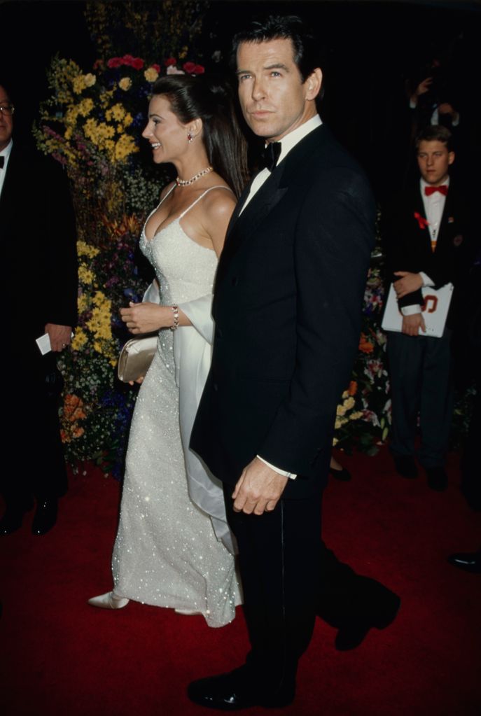 Keely Shaye Smith in a white sparkly dress and Pierce Brosnan in a suit with flowers in the background
