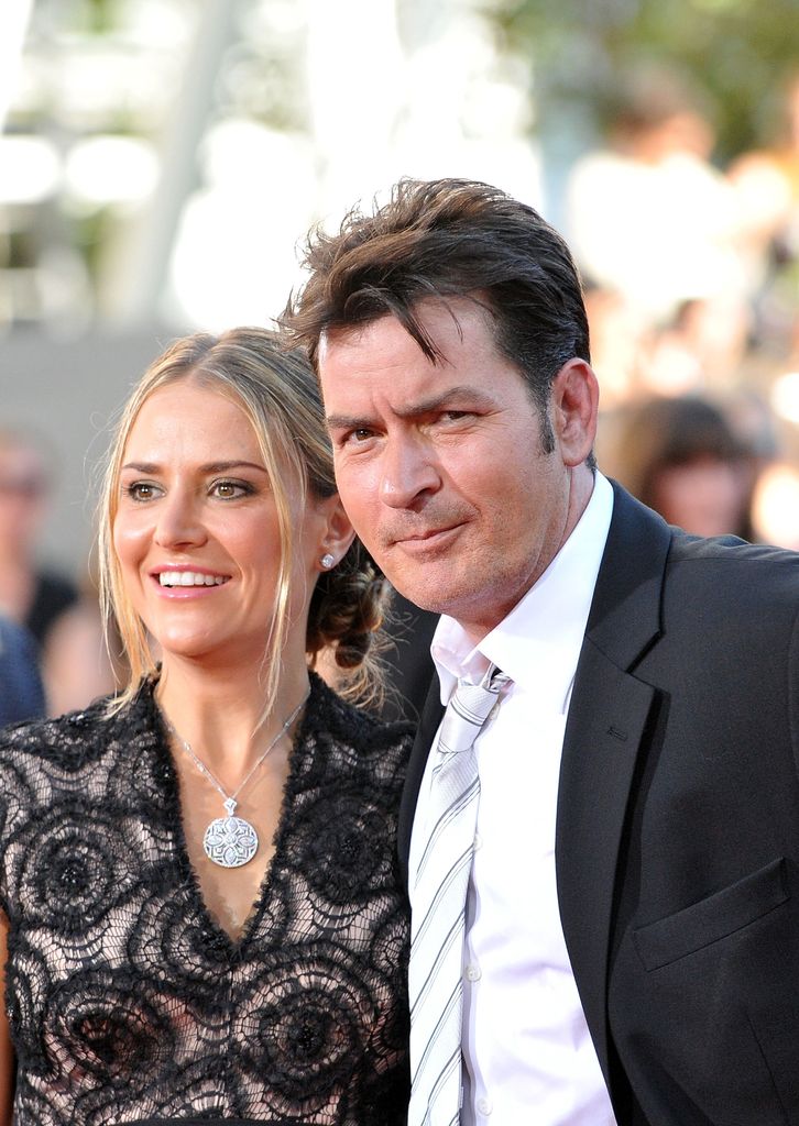 Charlie Sheen se tient devant sa femme Brooke Mueller. Il porte une veste noire avec une chemise blanche et une cravate. 
