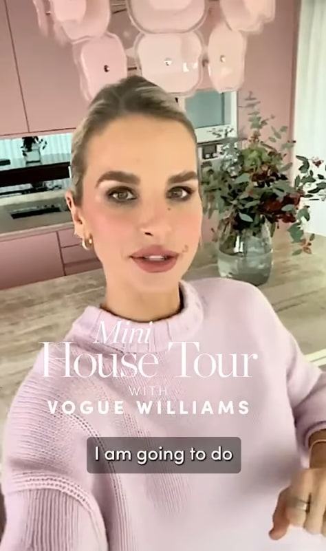 A woman in a light purple sweater stands in a modern kitchen with pink cabinets and a floral arrangement. Text on the image reads, Mini House Tour with Vogue Williams 