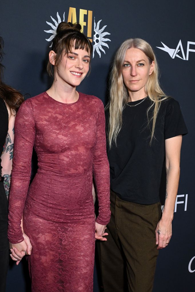 Kristen Stewart and Dylan Meyer  hold hands on the red carpet. Dylan wears a black t-shirt and khaki trousers