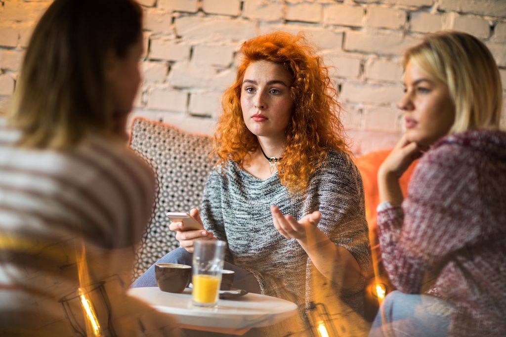 Worried redhead woman using cell phone while talking to her friends in a cafe