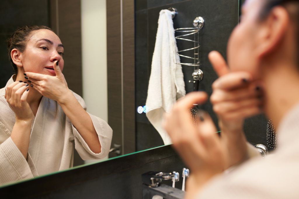 Woman in beige bathrobe looking at mirror and spreading moisturizing product on face during beauty routine in contemporary washroom.