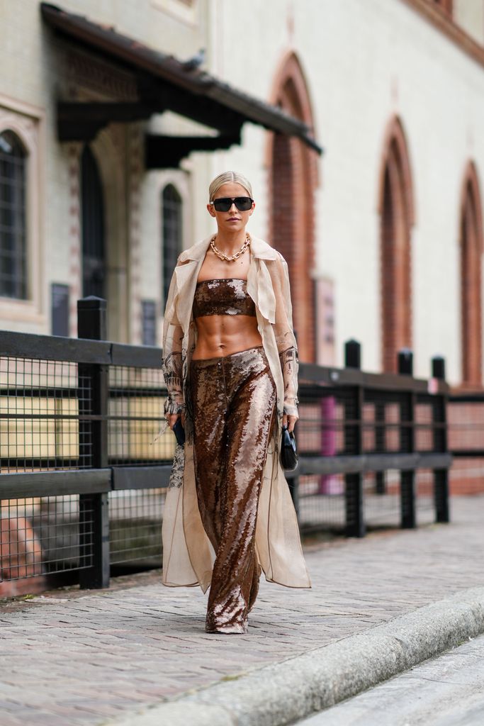 MILAN, ITALY - SEPTEMBER 20: Caroline Daur wears sunglasses, a golden chain necklace, a golden shiny sequined full outfit, bras / crop top, flared pants, a long white mesh jacket with printed features, outside Alberta Ferretti, during the Milan Fashion Week - Womenswear Spring/Summer 2024 on September 20, 2023 in Milan, Italy. (Photo by Edward Berthelot/Getty Images)