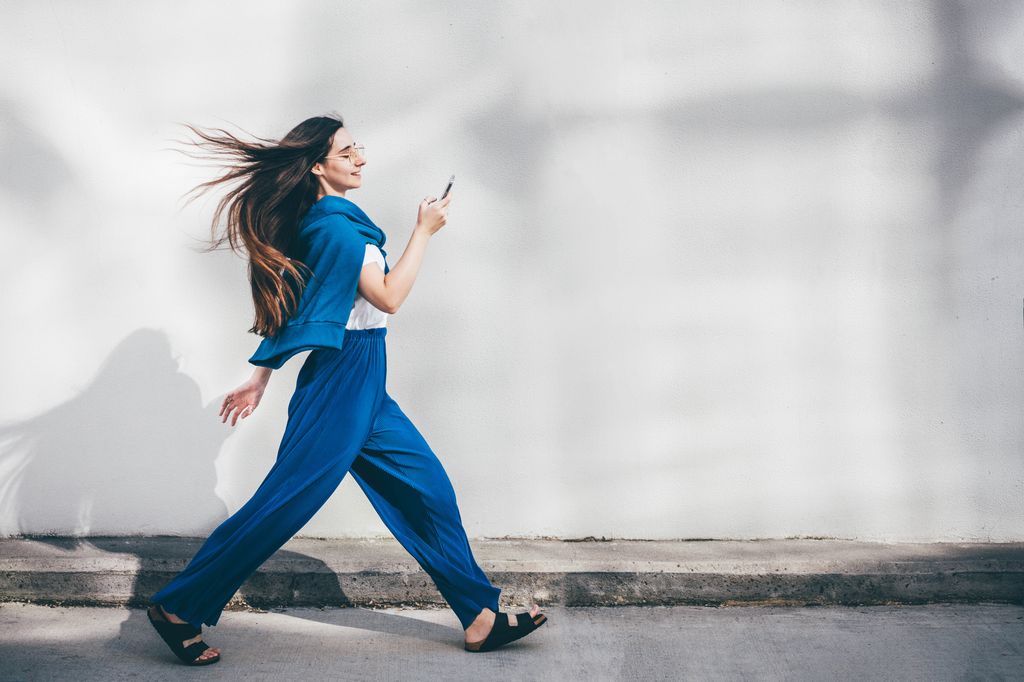 Woman using mobile phone while walking in front of concrete  wall.