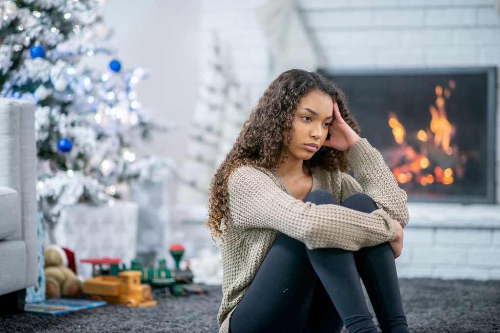 A beautiful girl of african descent sits on the carpet in her living room in front of the fireplace. She looks stressed and depressed as she sits alone on the holidays.