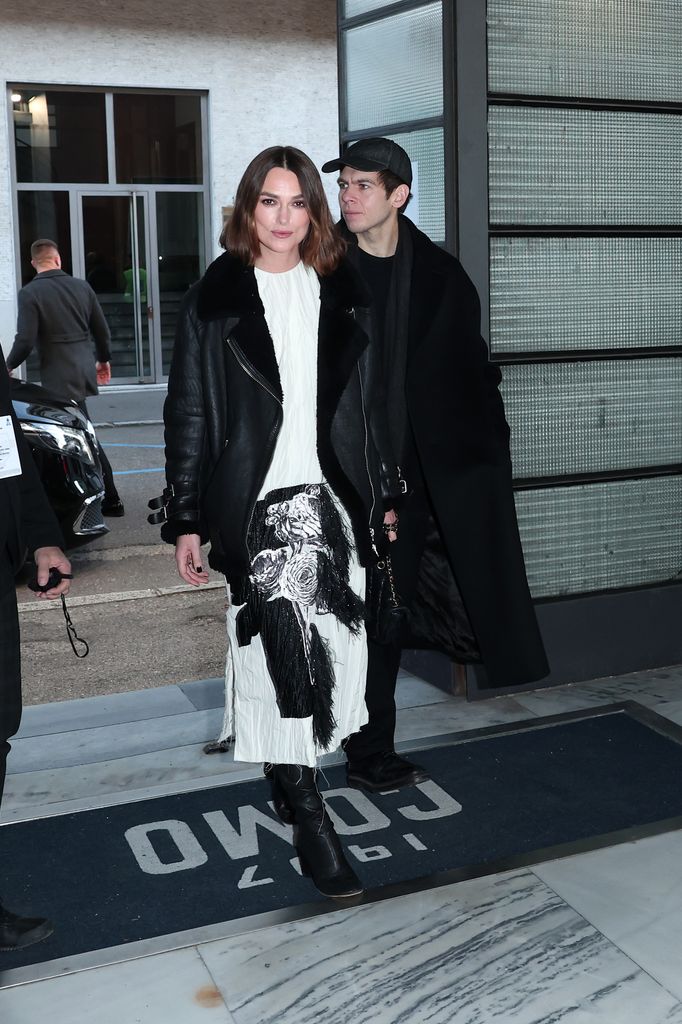 Keira Knightley steps forward confidently in a white midi dress with bold black floral embellishments. She pairs it with an oversized black shearling-lined leather jacket, black boots, and a sleek bob hairstyle. Beside her, James Righton wears a long black coat and cap.