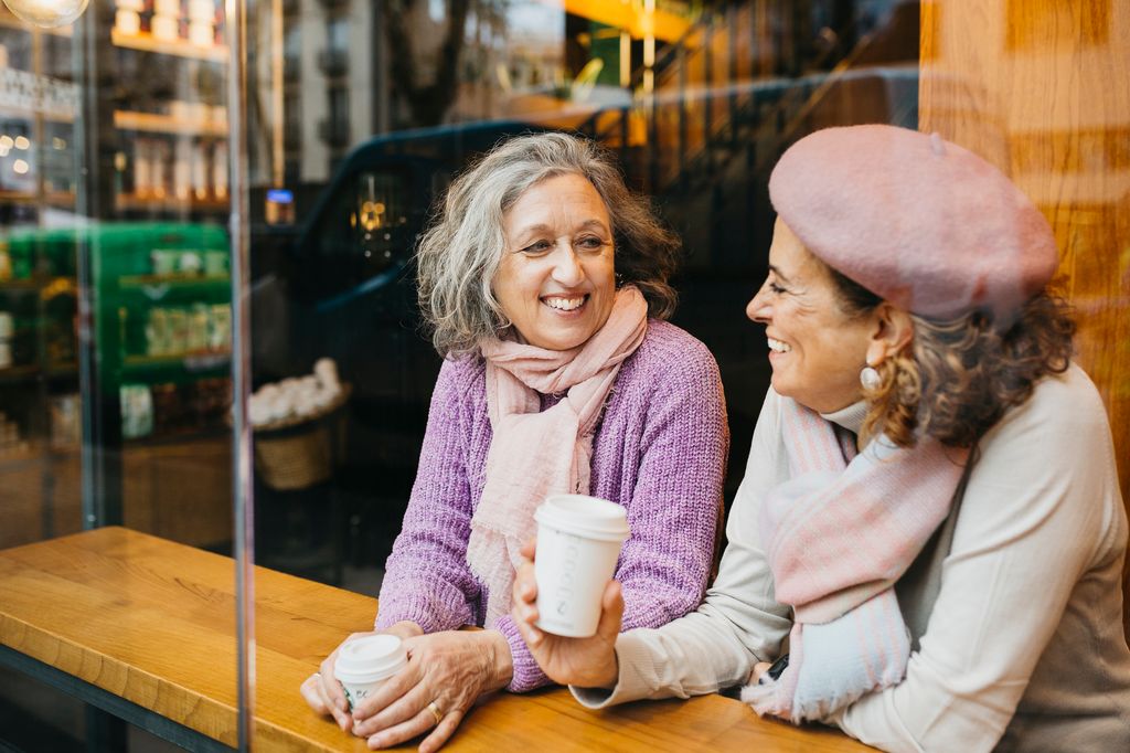 Two smiling senior women friends sitting in a coffee shop bar next to a window and drinking coffee