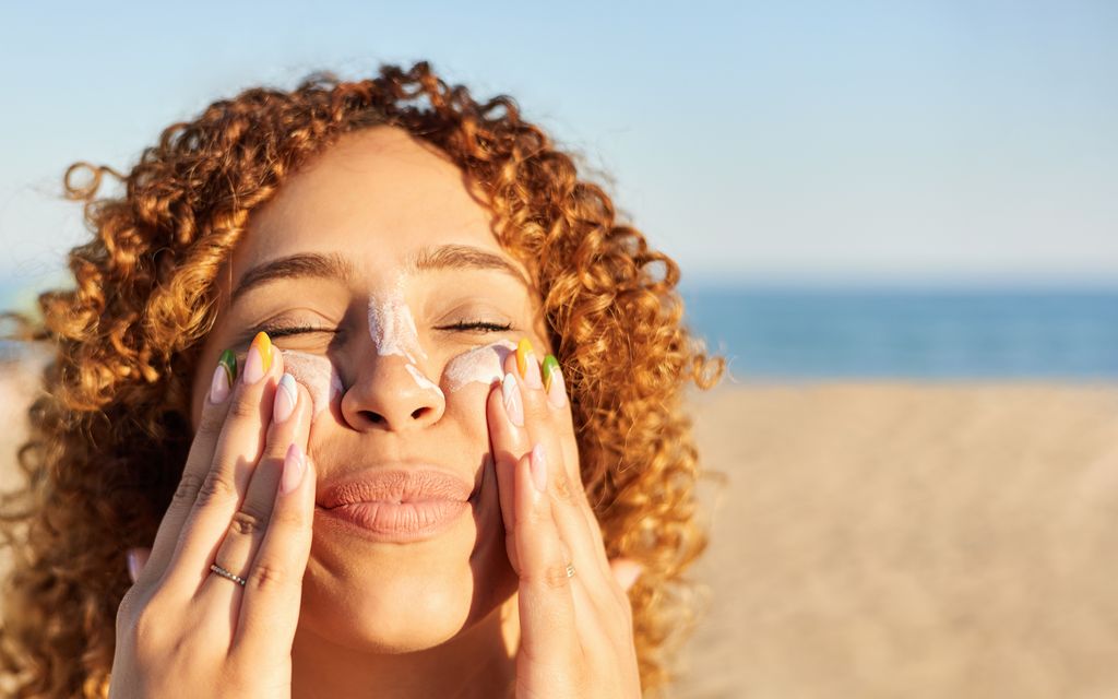 Young woman applying sunscreen to her face on the beach in a summer sunset.