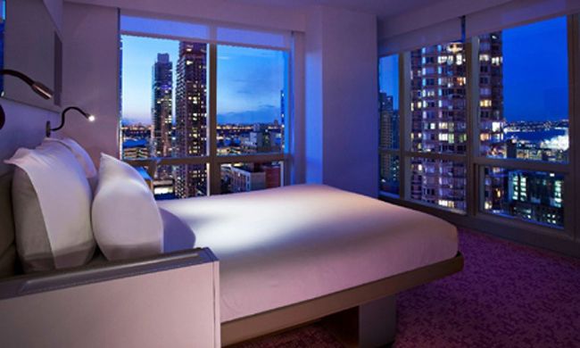 a bed in a room with a corner window boasting views of NY skyscrapers lit at night