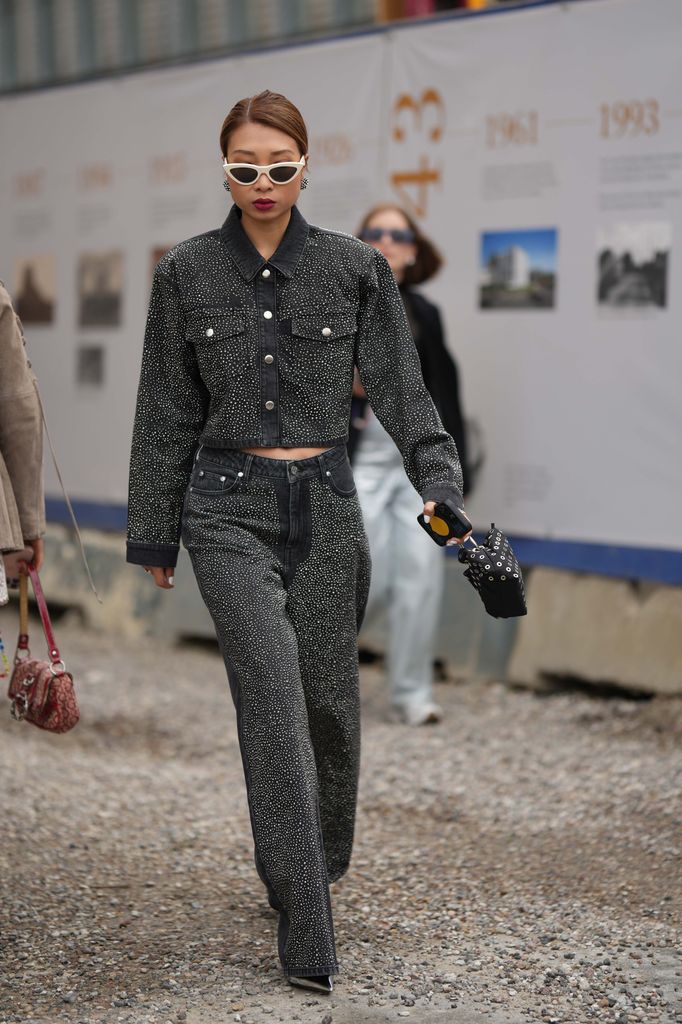 A guest wears white latte vintage sunglasses, black and crystals earrings, a dark gray denim with embroidered crystals buttoned jacket, matching dark gray denim with embroidered crystals wide legs pants, a black nylon nailed / studded handbag, 