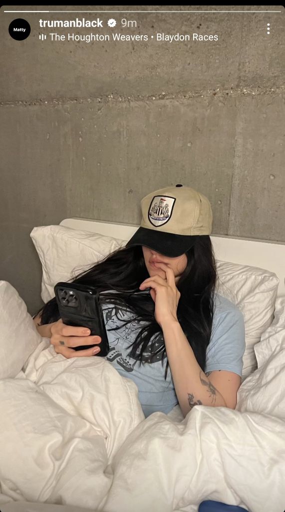 Woman in bed wearing a cap leaning against a concrete wall, snuggled in white duvets