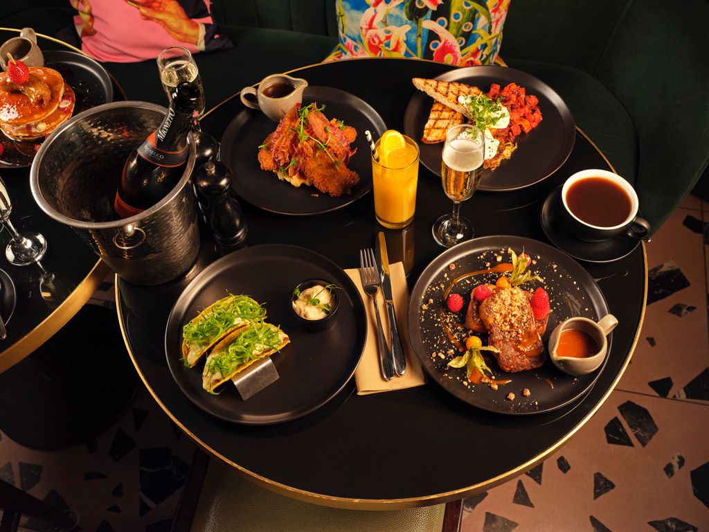 A top-down view of a stylish brunch spread at NXY London. Four black plates hold different dishes: pulled beef tacos with vibrant green garnish, fried chicken with a side, brioche French toast topped with tomatoes and a poached egg, and a sweet dish with berries and sauce. A bucket of chilled prosecco, two glasses of sparkling wine, a glass of orange juice, and a cup of coffee complete the table setting on a sleek black surface with gold trim.