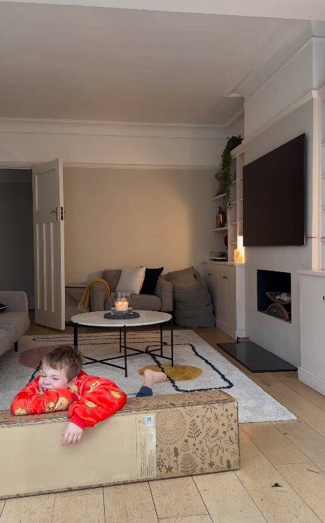 Danny Jones son lies on top of a box in their new living room with TV and sofas in the background