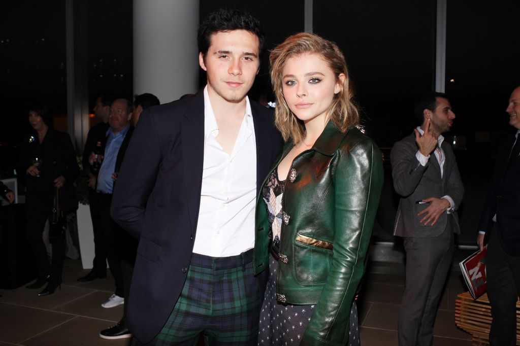 Brooklyn Beckham in tartan trousers and blazer posing with Chloe Grace Moretz who wears green leather jacket and polka dot skirt