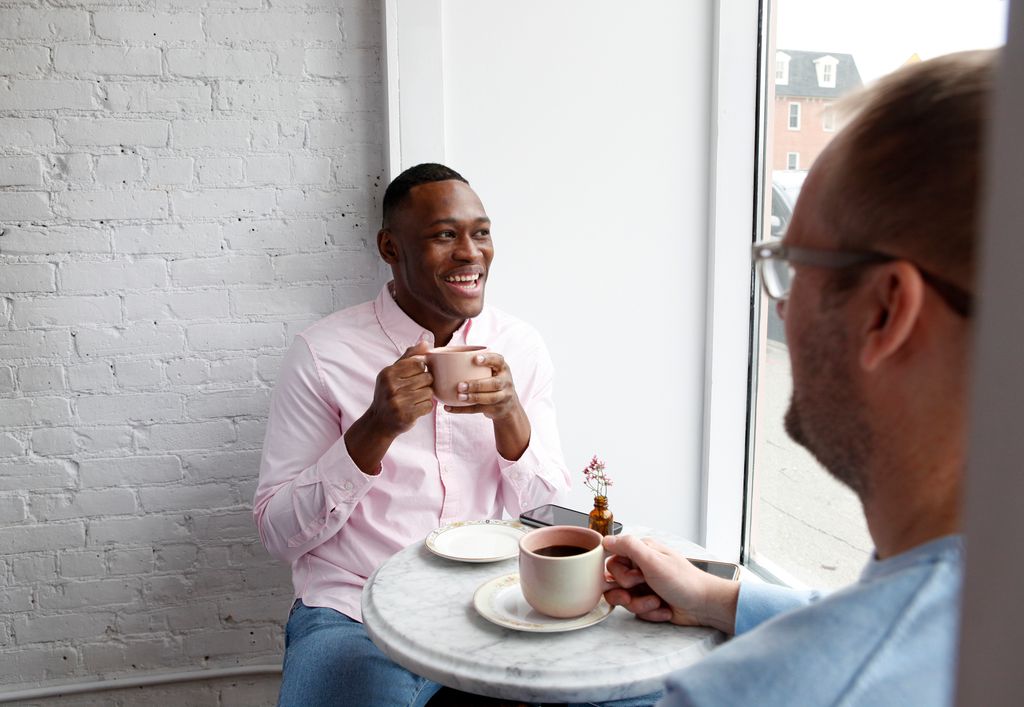 Man sitting at cafe table with a cup of coffee, smiling at another man