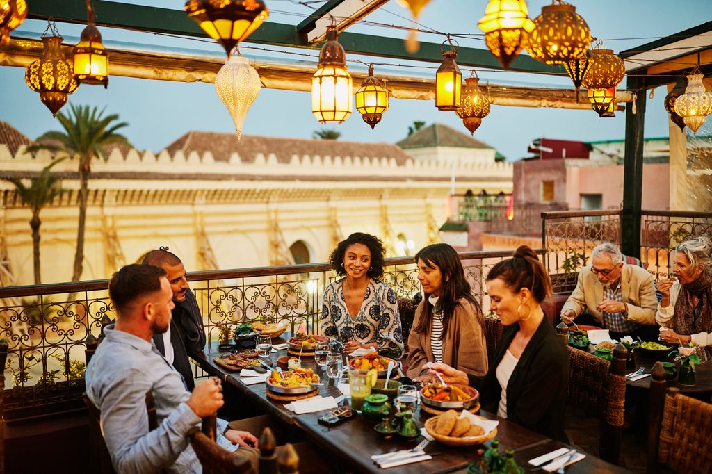 Wide shot of tourists laughing and savoring traditional Moroccan tajine at scenic rooftop eatery during holiday in Marrakech at night