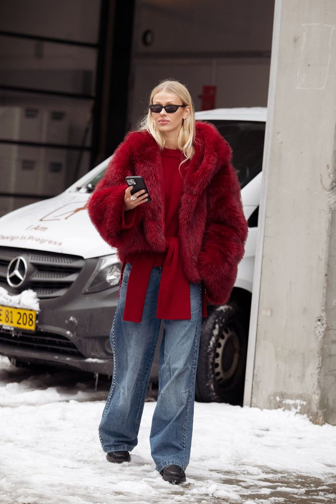 A guest wears blue wide jeans, red sweater, red fur coat and sunglasses outside the Forza Collective show during Copenhagen Fashion Week AW26