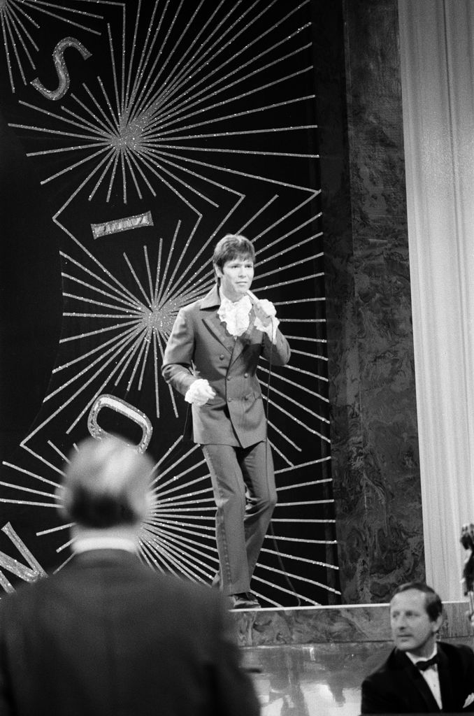 Cliff Richard performs at Eurovision 1968 in a double-breasted suit with dramatic ruffled cuffs, channeling classic 60s British pop style.