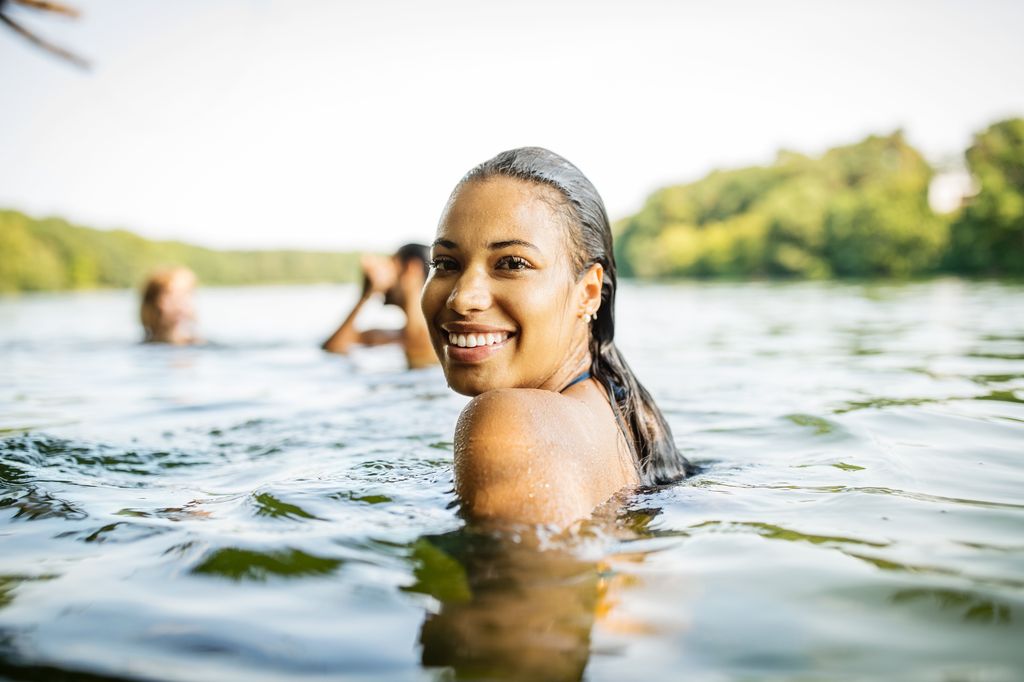 Smiling young woman in a lake with friends swimming in the background. Beautiful woman swimming in lake and looking at camera.
