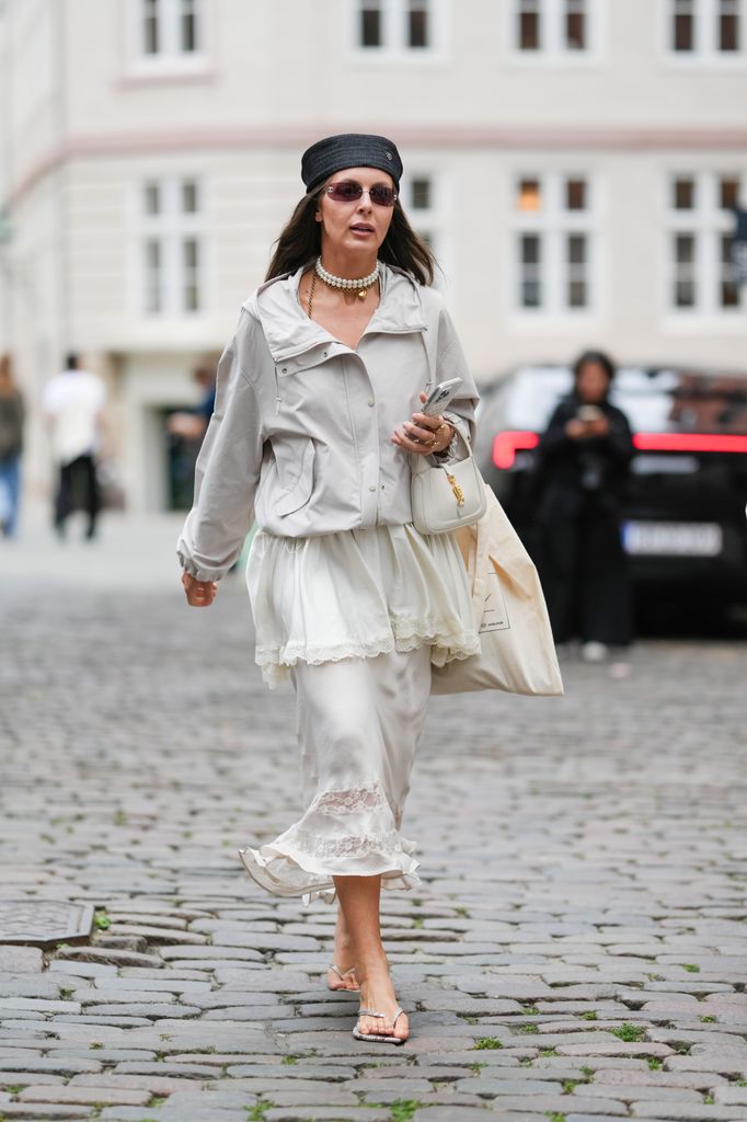 A guest wears long straight dark brown hair under a wide black headband, narrow oval sunglasses with brown-tinted lenses, a multi-strand pearl choker layered with a delicate gold necklace with a small pendant, and a gold bracelet with rings; a light gray hooded zip-front jacket with a snap placket and side pockets; 
