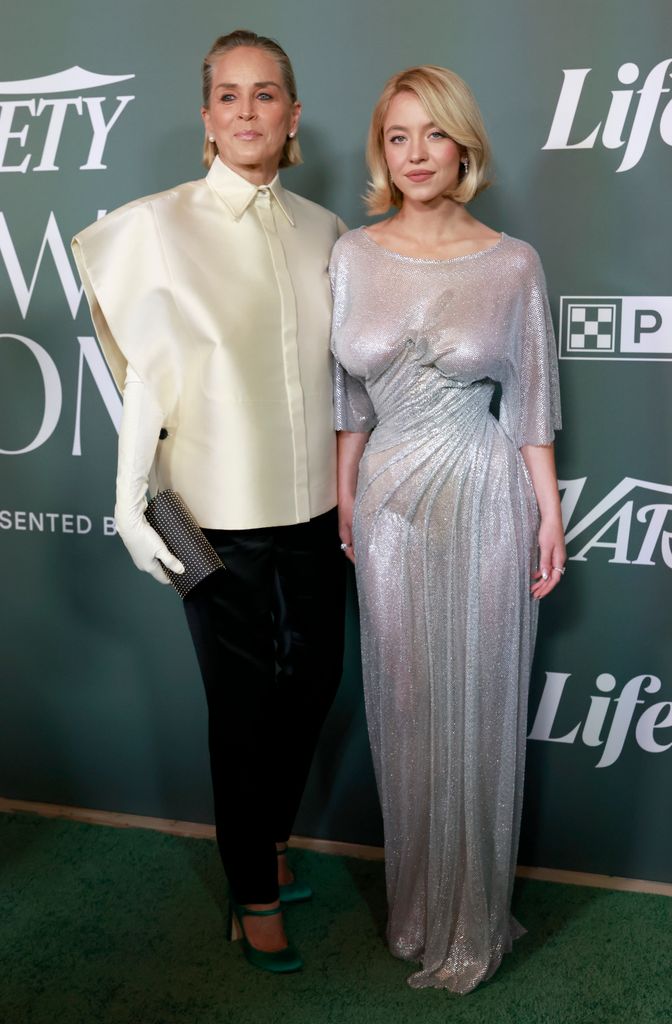 Sharon Stone and Sydney Sweeney stand side by side on the red carpet. Sharon wears an oversize cream shirt with short sleeves, teamed with long white gloves, black trousers and black heels.