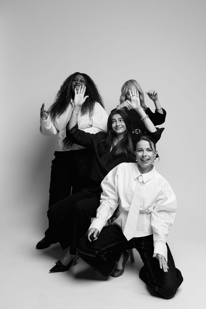Playful black-and-white photo of four women in tailored outfits laughing and raising their hands in a candid studio moment.