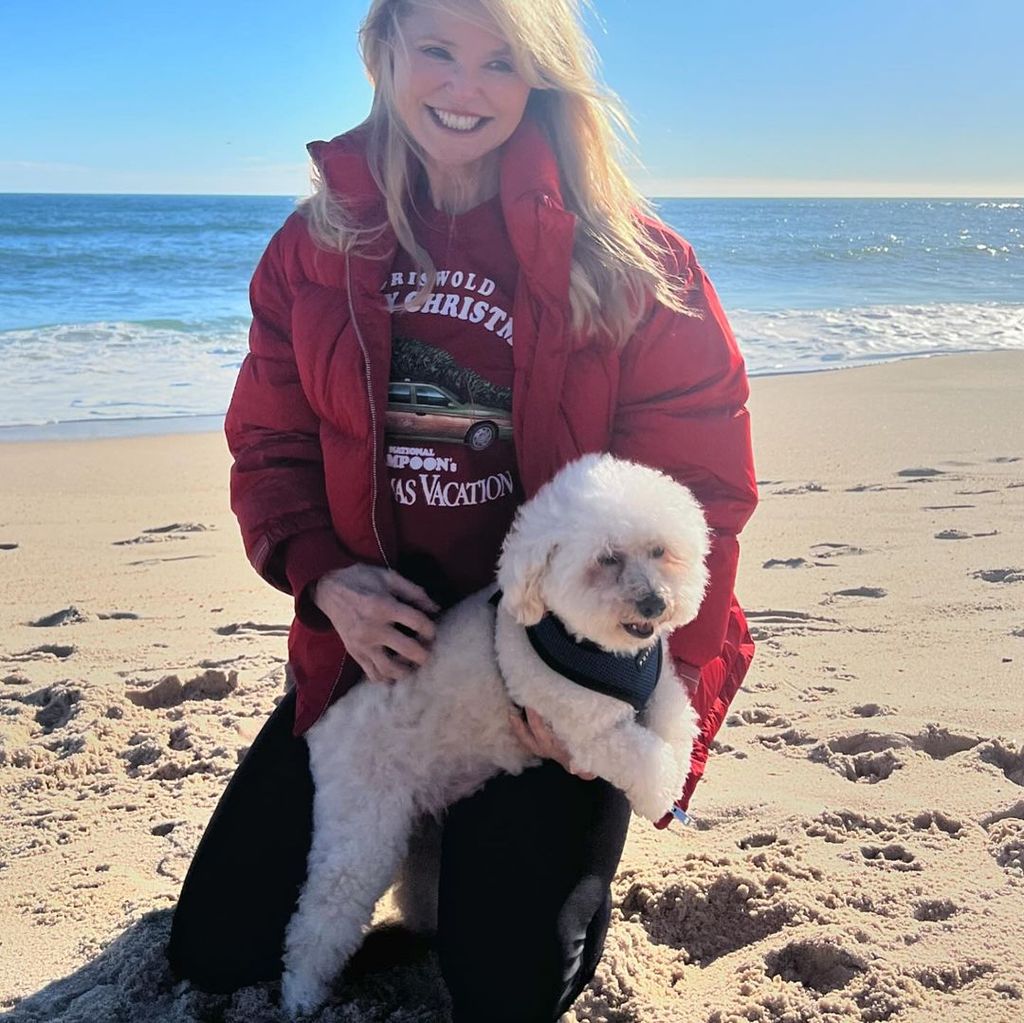Christie Brinkley knees in sand with her dog on her lap