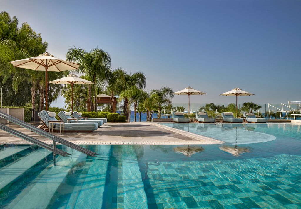 Blue pool with steps going in, surrounded by sun loungers, umbrellas and palm trees