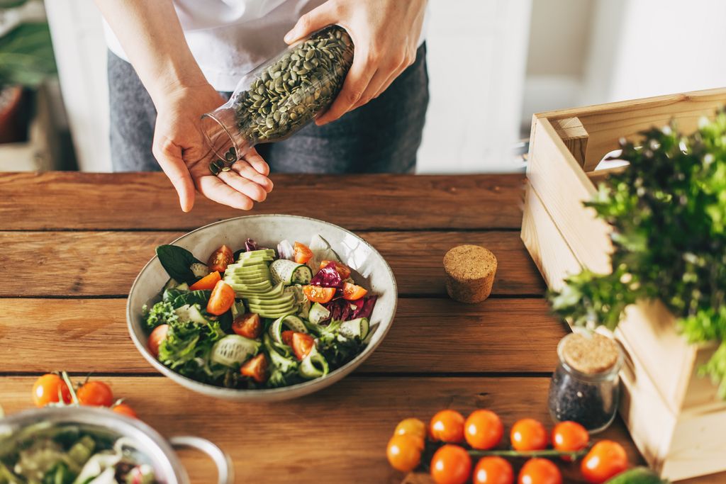 Close Up Of Woman Adding Pumpkin Seeds To Healthy Vegan Meal In Bowl