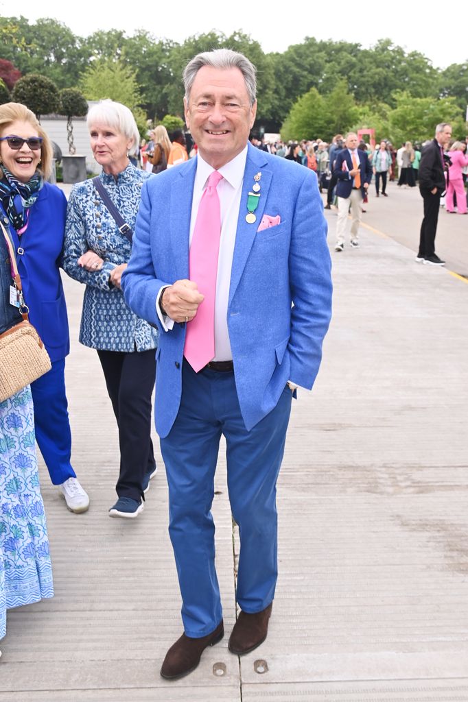 Alan Titchmarsh in a bright blue blazer, pink tie and medals poses at the Chelsea Flower Show surrounded by other attendees.
