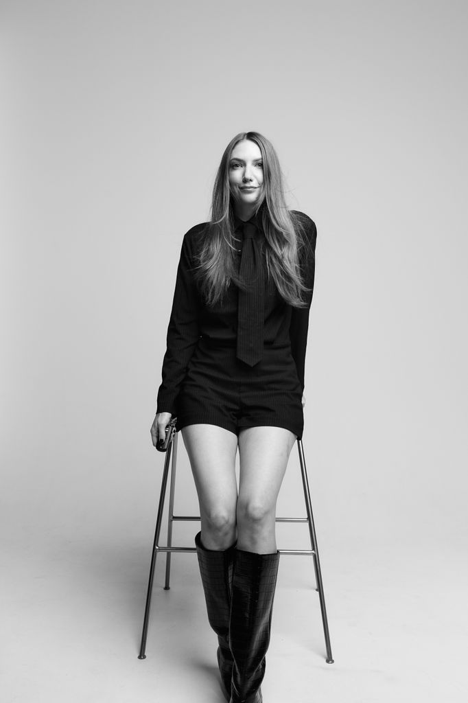 Black-and-white studio portrait of a woman seated on a high stool wearing an all-black outfit with long sleeves and knee-high textured boots.