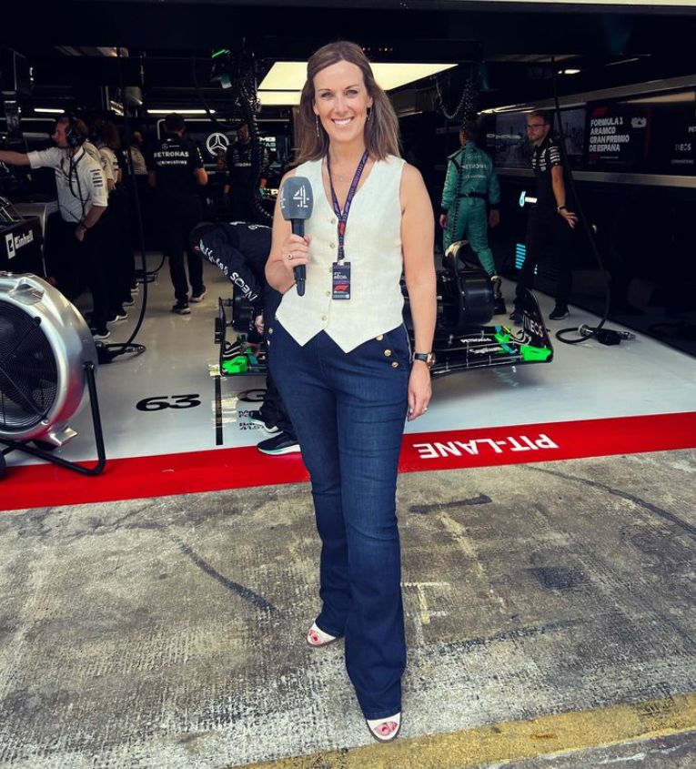 Lee McKenzie holding a microphone in front of an F1 garage