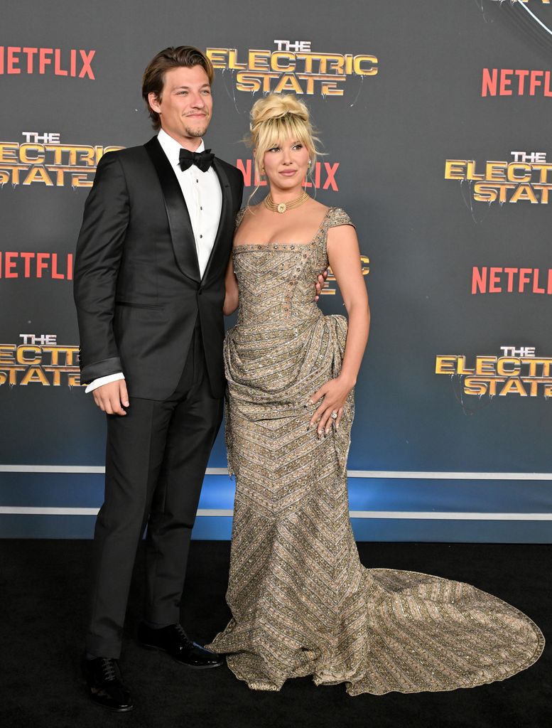 Millie Bobby Brown poses alongside a man in a classic black tuxedo with a white shirt and black bow tie. She is wearing the same embellished gown, with her hand resting on her hip. The man smiles beside her, his arm wrapped around her waist. The backdrop remains the same, with the "Netflix" and "The Electric State" branding visible.