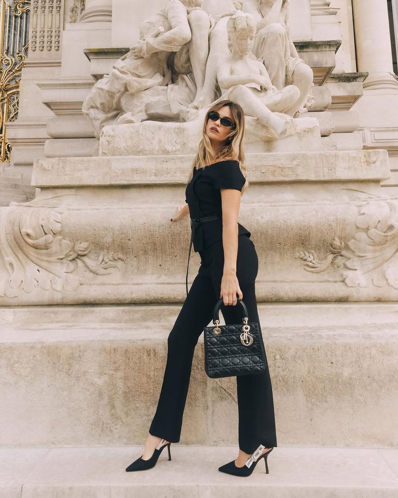 Lily James poses confidently outside the Petit Palais in Paris during Fashion Week, wearing black Dior trousers and heels with a Lady Dior bag. She stands beside an elaborate marble sculpture, exuding effortless French girl elegance.