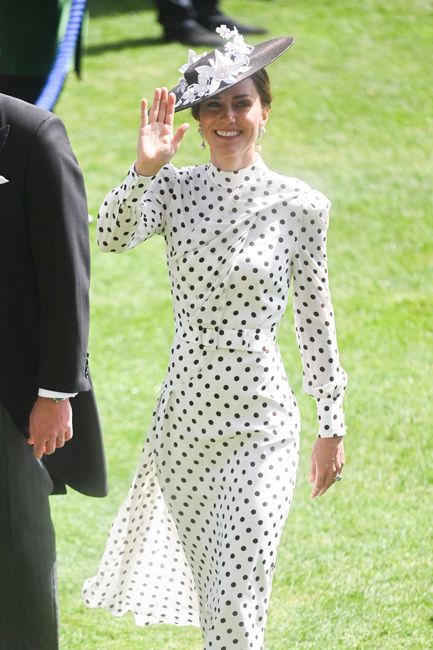 kate waves her hand and waves while wearing a form fitting white dress with long sleeves a nipped in waist and a floaty sheer skirt overlay and the dress is covered in black dots which coordinate with her black hat that is decorated with white flowers