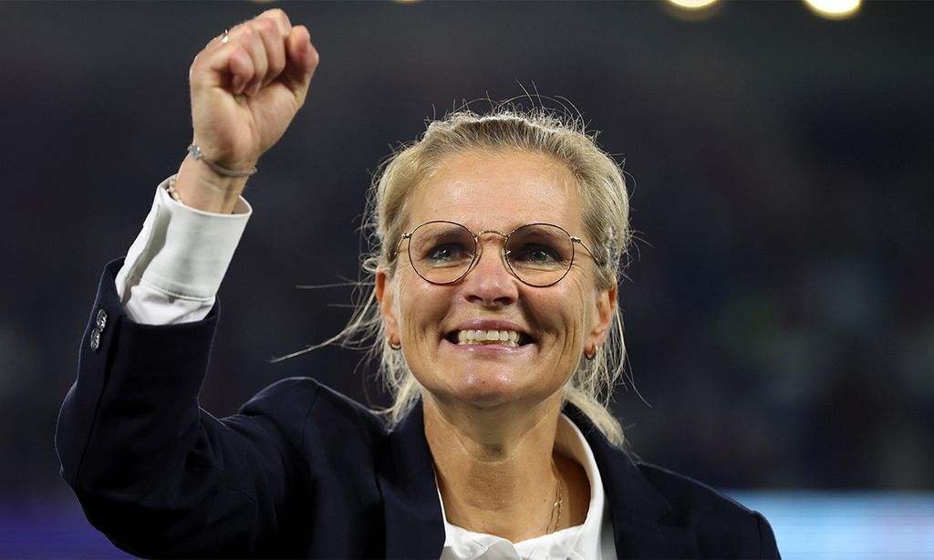 Sarina Wiegman wearing glasses and a dark jacket smiles and raises a clenched fist