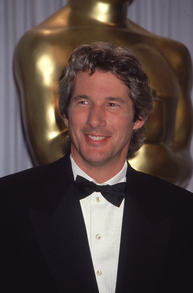 Richard Gere, wearing a tuxedo, standing and smiling in front of a gold, life size statue of the Oscar, symbol of the Academy of Motion Arts and Sciences, at an Academy Awards ceremony, California, 1993