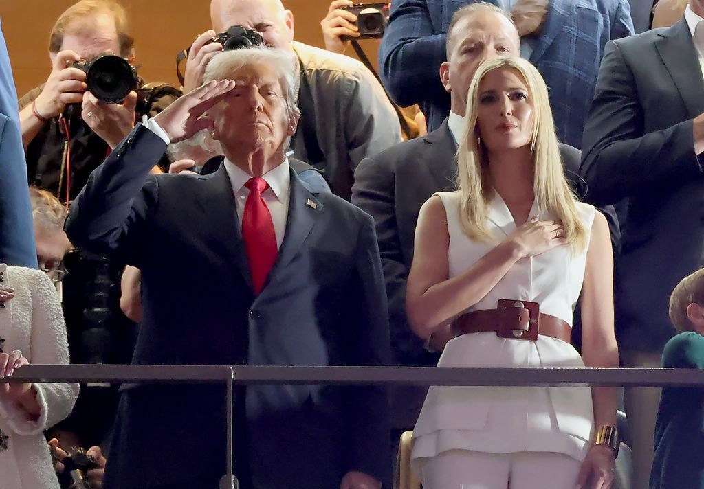 Ivanka Trump stands in a stadium suite, wearing a white sleeveless vest with matching trousers and a brown belt, her blonde hair sleek as she places a hand over her heart.