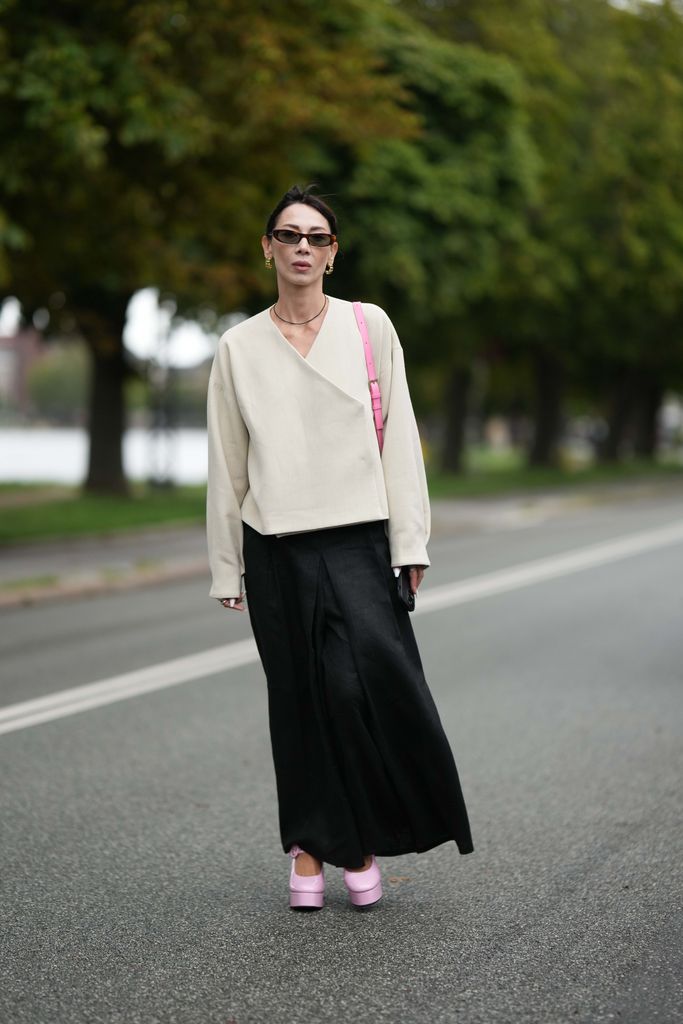 A guest wears brown sunglasses, gold earrings, a black small pearls necklace, a white latte wrap jacket, a pale pink shiny leather shoulder bag, a black silk long dress, a pale pink shiny leather varnished platform soles high heels shoes , outside Stine Goya, during the Copenhagen Fashion Week Spring/Summer 2024 on August 09, 2023 in Copenhagen, Denmark. (Photo by Edward Berthelot/Getty Images)